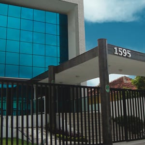 No suntuoso prédio espelhado se encontra o escritório de Recife/PE, protegido por uma extensa grade preta e câmera de segurança.