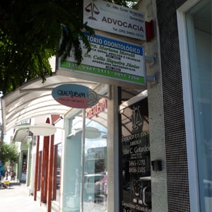 Entre o comércio de roupas, em Monte Sião/MG, nota-se o escritório de advocacia na estreita porta de vidro preto.